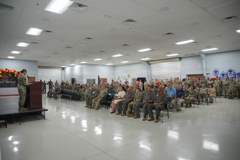 A military ceremony or briefing is taking place in a large indoor facility, likely on an Air Force base. A uniformed speaker stands at a podium on a raised stage addressing a large audience of military personnel and some civilians seated in rows of chairs. A banner on the podium reads '2d Wing,' indicating this is associated with the 2nd Wing unit.