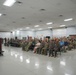 A military ceremony or briefing is taking place in a large indoor facility, likely on an Air Force base. A uniformed speaker stands at a podium on a raised stage addressing a large audience of military personnel and some civilians seated in rows of chairs. A banner on the podium reads '2d Wing,' indicating this is associated with the 2nd Wing unit.