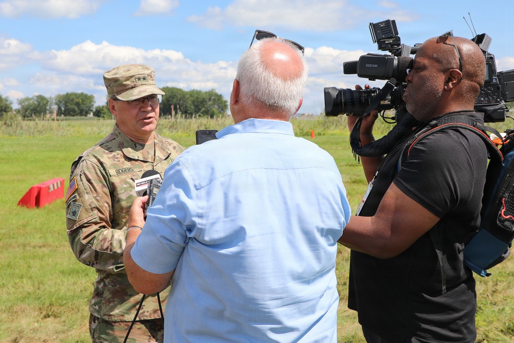 A U.S. Army officer wearing camouflage uniform with the name tape 'ROGERS' and two-star general rank insignia is being interviewed by a television news crew outdoors. A reporter holding a microphone faces the general while a cameraman operates a professional video camera. The setting appears to be an open field or training area on a sunny day.