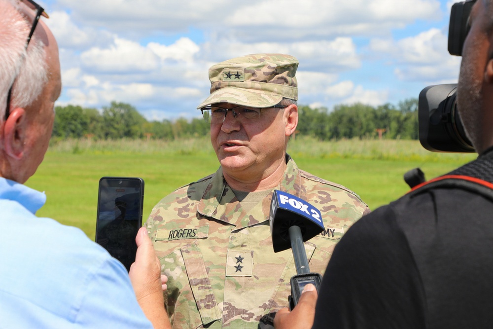A U.S. Army Major General named Rogers is conducting a media interview outdoors, speaking to reporters from FOX 2 and at least one other journalist. He is wearing an OCP (Operational Camouflage Pattern) uniform with two-star rank insignia visible on both his cap and chest. The setting appears to be a grassy field or military installation on a partly cloudy day.