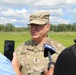 A U.S. Army Major General named Rogers is conducting a media interview outdoors, speaking to reporters from FOX 2 and at least one other journalist. He is wearing an OCP (Operational Camouflage Pattern) uniform with two-star rank insignia visible on both his cap and chest. The setting appears to be a grassy field or military installation on a partly cloudy day.