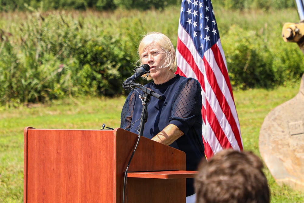 A woman with blonde hair is speaking at a wooden podium equipped with a microphone at an outdoor event. An American flag is displayed prominently behind her, and construction equipment is partially visible in the background. The setting and equipment suggest this may be a groundbreaking ceremony or official outdoor event.