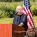 A woman with blonde hair is speaking at a wooden podium equipped with a microphone at an outdoor event. An American flag is displayed prominently behind her, and construction equipment is partially visible in the background. The setting and equipment suggest this may be a groundbreaking ceremony or official outdoor event.