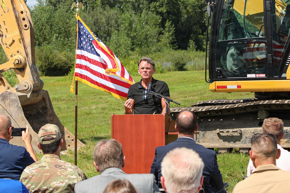 An outdoor groundbreaking ceremony is taking place, with a man in a black polo shirt speaking at a wooden podium. An American flag with gold fringe stands prominently to his left, flanked by two yellow CAT excavators. An audience of attendees, including at least one person in military camouflage uniform, faces the speaker.