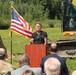 An outdoor groundbreaking ceremony is taking place, with a man in a black polo shirt speaking at a wooden podium. An American flag with gold fringe stands prominently to his left, flanked by two yellow CAT excavators. An audience of attendees, including at least one person in military camouflage uniform, faces the speaker.