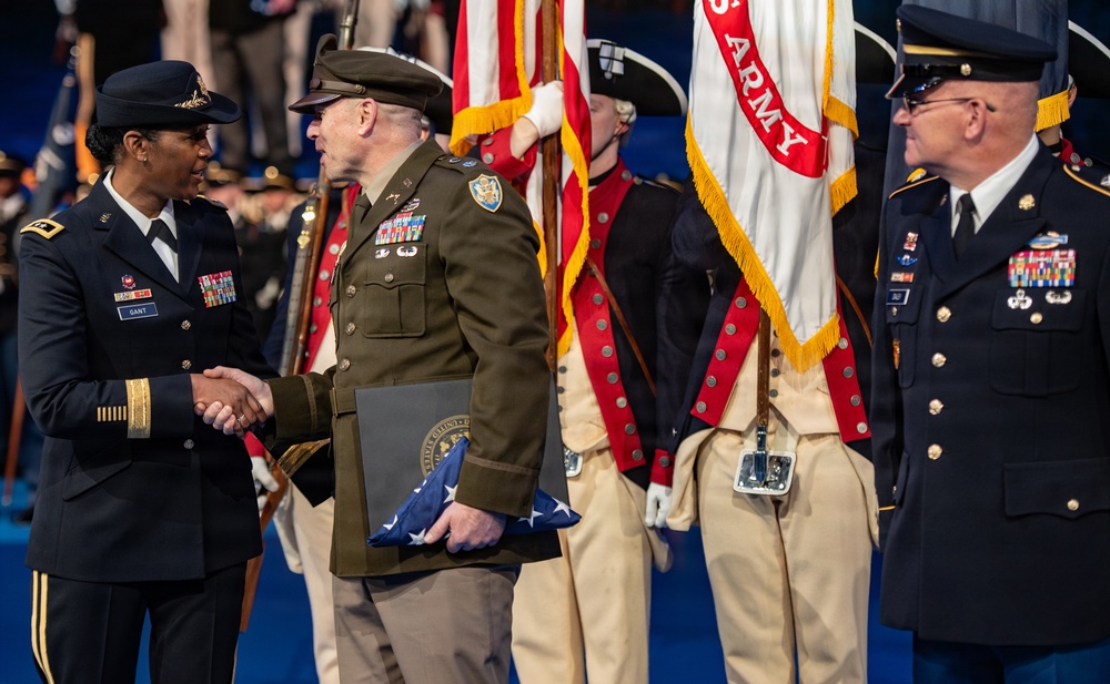 A formal U.S. Army military ceremony is taking place, likely a retirement or change of command event. A female Army officer with the name tag 'GANT' is shaking hands with a senior male officer in an olive dress uniform who is holding a folded American flag and a certificate folder. Color guard soldiers in historical Revolutionary War-era uniforms stand in the background alongside a U.S. Army flag.