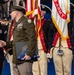 A formal U.S. Army military ceremony is taking place, likely a retirement or change of command event. A female Army officer with the name tag 'GANT' is shaking hands with a senior male officer in an olive dress uniform who is holding a folded American flag and a certificate folder. Color guard soldiers in historical Revolutionary War-era uniforms stand in the background alongside a U.S. Army flag.