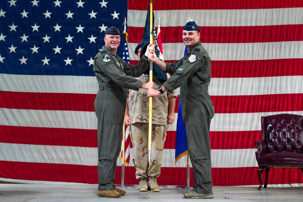 Two U.S. Air Force officers in olive green flight suits are participating in a formal change of command ceremony, passing a unit guidon (flag) between them. The ceremony takes place in front of a large American flag backdrop, with a third service member in desert camouflage uniform visible in the background. A burgundy leather chair is positioned to the right side of the stage.