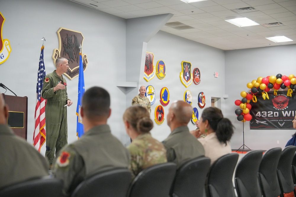 A military ceremony or briefing is taking place at Creech Air Force Base, Nevada, associated with the 432d Wing. A uniformed officer in a green flight suit is addressing an audience of seated military personnel and civilians. The room is decorated with unit patches/emblems on the walls, an American flag, an Air Force flag, and a balloon arch in red, black, and gold colors.