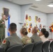 A military ceremony or briefing is taking place at Creech Air Force Base, Nevada, associated with the 432d Wing. A uniformed officer in a green flight suit is addressing an audience of seated military personnel and civilians. The room is decorated with unit patches/emblems on the walls, an American flag, an Air Force flag, and a balloon arch in red, black, and gold colors.