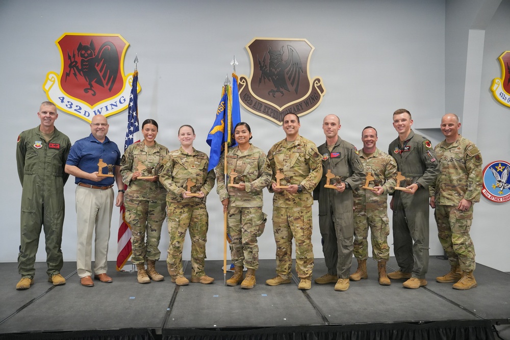 A group of approximately ten military personnel and one civilian pose for a formal awards photo on a stage. Most individuals are holding wooden trophy/award plaques, suggesting this is a recognition or awards ceremony. The backdrop features unit insignia shields for the 432nd Wing and 432nd Air Expeditionary Wing, along with an American flag and a military guidon.