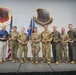 A group of approximately ten military personnel and one civilian pose for a formal awards photo on a stage. Most individuals are holding wooden trophy/award plaques, suggesting this is a recognition or awards ceremony. The backdrop features unit insignia shields for the 432nd Wing and 432nd Air Expeditionary Wing, along with an American flag and a military guidon.
