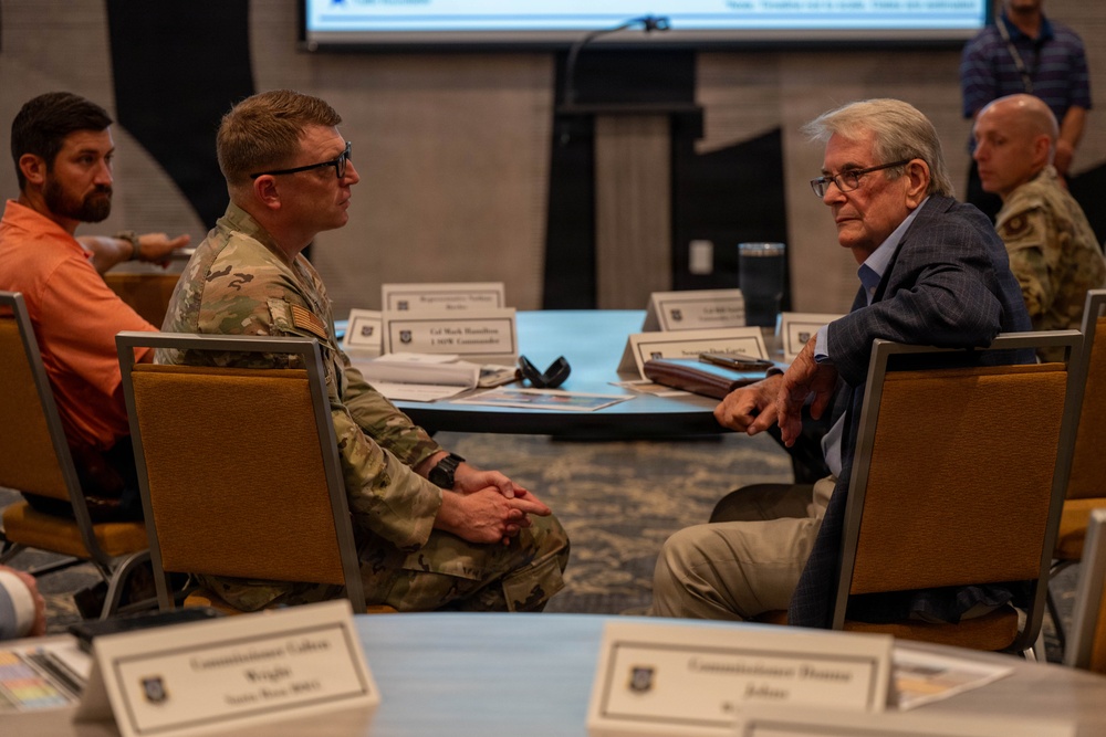 A formal meeting or briefing is taking place at a conference table, with military personnel in camouflage uniforms and civilians in business attire seated across from each other. Name placards are visible on the table, indicating an organized, official gathering. A presentation screen is visible in the background, suggesting a structured briefing or coordination session.