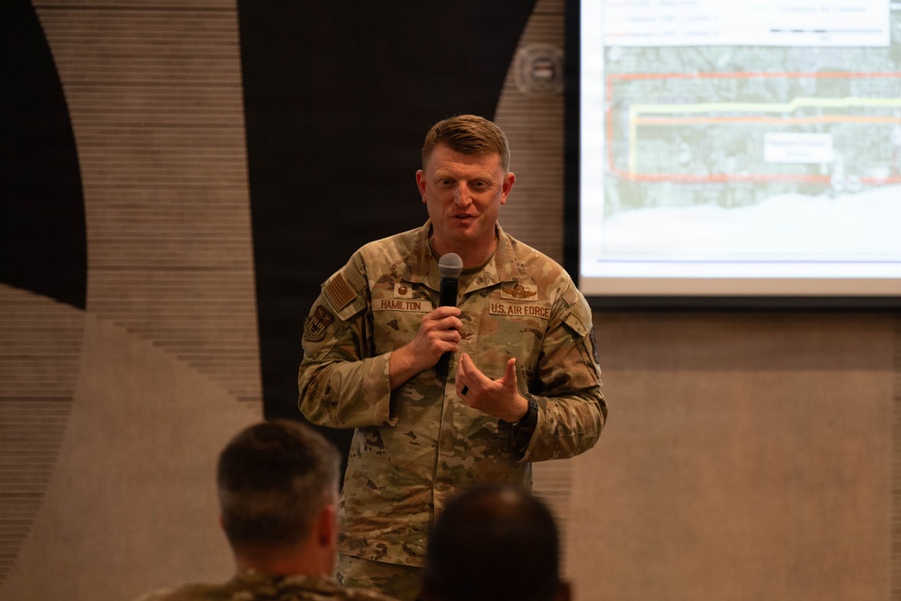 A U.S. Air Force servicemember named Hamilton is delivering a briefing or presentation while holding a microphone. He is wearing OCP (Operational Camouflage Pattern) uniform with 'U.S. AIR FORCE' and 'HAMILTON' name tape visible. A projected screen displaying what appears to be an aerial map or facility diagram is visible in the background.