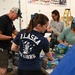A group of volunteers are working together at a table sorting and packing food and supplies, likely at a food bank or community relief distribution center. One volunteer is prominently wearing an 'Alaska Air Guard' t-shirt. The setting appears to be a garage or warehouse space decorated with a birthday banner.