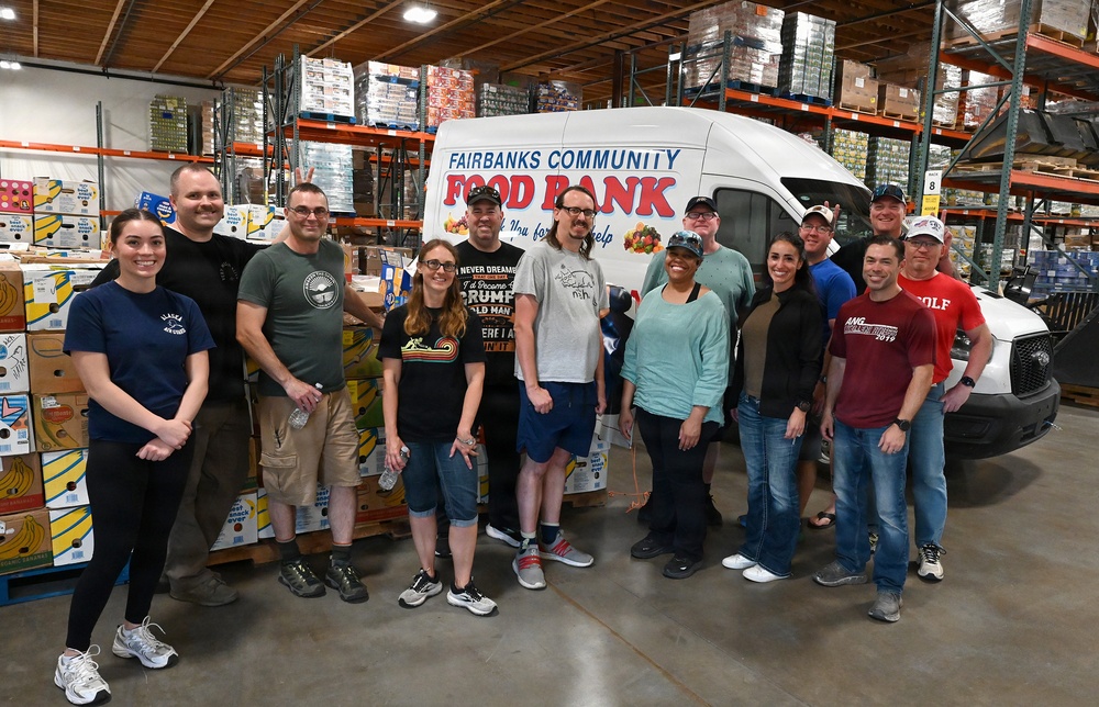 A group of approximately 15 volunteers poses for a group photo inside a warehouse facility for the Fairbanks Community Food Bank. They are standing in front of a white delivery van branded with the food bank's name and logo. The warehouse is stocked with pallets of food and supplies on industrial shelving racks.