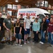 A group of approximately 15 volunteers poses for a group photo inside a warehouse facility for the Fairbanks Community Food Bank. They are standing in front of a white delivery van branded with the food bank's name and logo. The warehouse is stocked with pallets of food and supplies on industrial shelving racks.