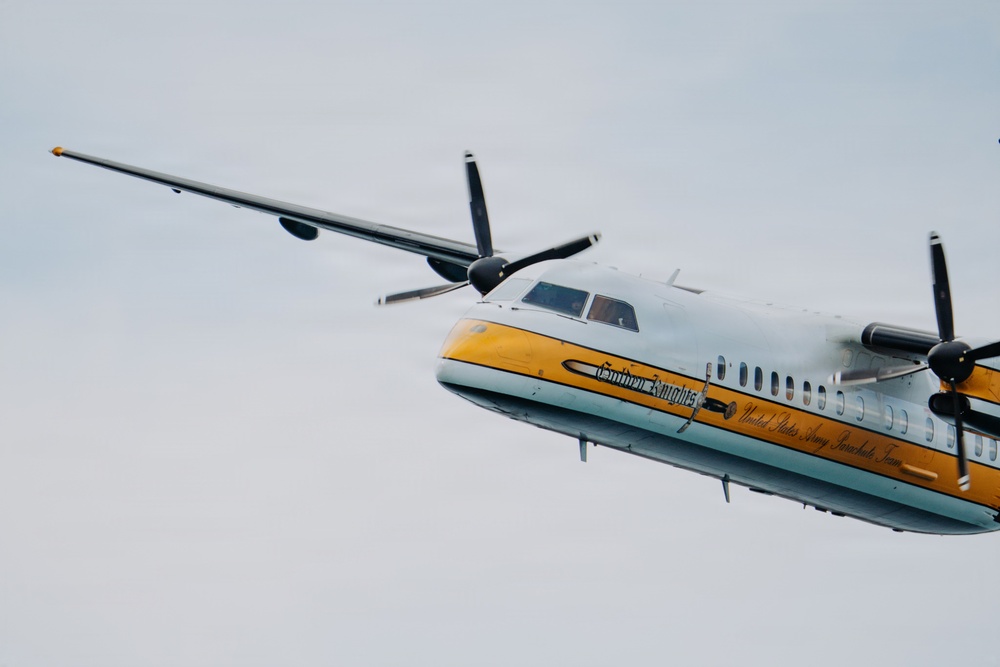 A twin-turboprop aircraft belonging to the United States Army Parachute Team, known as the 'Golden Knights,' is photographed in flight against an overcast sky. The aircraft features a distinctive white and yellow livery with 'Golden Knights' and 'United States Army Parachute Team' text visible on the fuselage. The image captures the aircraft at a slight angle, showing both propeller engines and the full wing span.