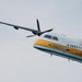 A twin-turboprop aircraft belonging to the United States Army Parachute Team, known as the 'Golden Knights,' is photographed in flight against an overcast sky. The aircraft features a distinctive white and yellow livery with 'Golden Knights' and 'United States Army Parachute Team' text visible on the fuselage. The image captures the aircraft at a slight angle, showing both propeller engines and the full wing span.