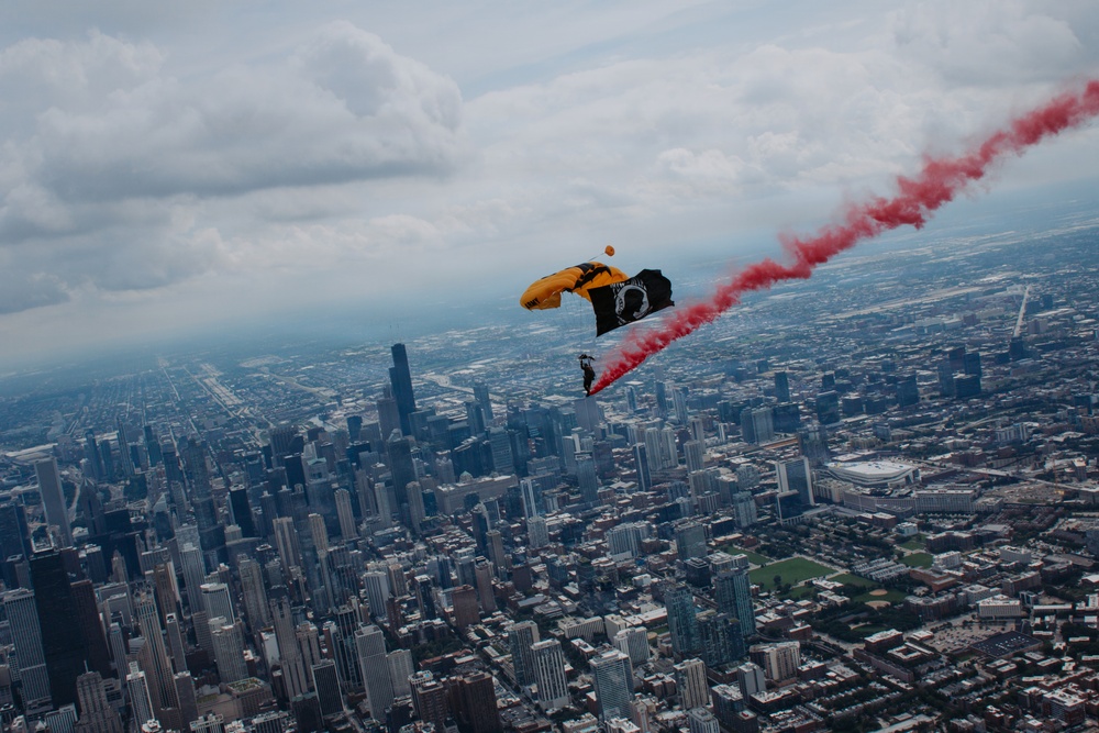 A skydiver with a yellow parachute descends over a major urban skyline, trailing red smoke and carrying a black POW/MIA flag. The aerial perspective reveals a dense cityscape consistent with Chicago, Illinois, including recognizable skyscrapers and urban grid patterns. The scene appears to be part of a ceremonial or airshow demonstration.