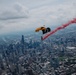A skydiver with a yellow parachute descends over a major urban skyline, trailing red smoke and carrying a black POW/MIA flag. The aerial perspective reveals a dense cityscape consistent with Chicago, Illinois, including recognizable skyscrapers and urban grid patterns. The scene appears to be part of a ceremonial or airshow demonstration.