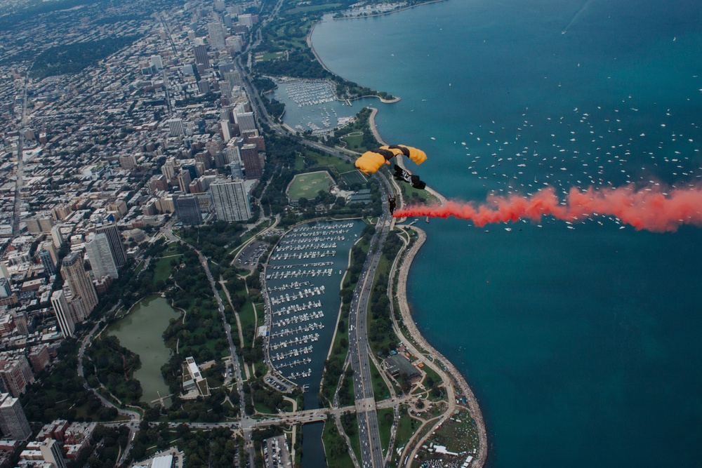 An aerial photograph captures a skydiver with a yellow parachute deploying red smoke as they descend over a major lakefront city, identifiable by its dense urban grid, green parkland, and large marina along the shoreline. The image is taken from a high altitude, providing a sweeping bird's-eye view of the cityscape, Lake Michigan, and numerous boats on the water. The red smoke trail creates a dramatic visual contrast against the blue lake and urban landscape below.