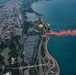 An aerial photograph captures a skydiver with a yellow parachute deploying red smoke as they descend over a major lakefront city, identifiable by its dense urban grid, green parkland, and large marina along the shoreline. The image is taken from a high altitude, providing a sweeping bird's-eye view of the cityscape, Lake Michigan, and numerous boats on the water. The red smoke trail creates a dramatic visual contrast against the blue lake and urban landscape below.