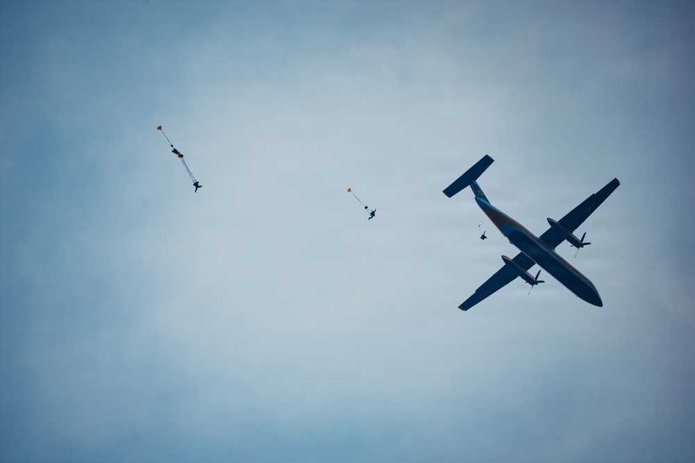 A turboprop aircraft is captured in flight against an overcast blue-grey sky, with multiple skydivers visible at various stages of descent. At least three skydivers can be seen, with two having deployed small pilot/drogue chutes visible in orange, and one appearing to still be near the aircraft. The scene depicts an active skydiving operation with jumpers having recently exited the plane.