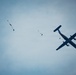A turboprop aircraft is captured in flight against an overcast blue-grey sky, with multiple skydivers visible at various stages of descent. At least three skydivers can be seen, with two having deployed small pilot/drogue chutes visible in orange, and one appearing to still be near the aircraft. The scene depicts an active skydiving operation with jumpers having recently exited the plane.