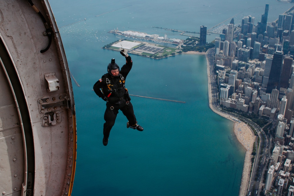 A skydiver in a black jumpsuit and helmet is captured mid-exit from an aircraft, with one arm raised, against a stunning aerial backdrop of Chicago's lakefront. The image shows Lake Michigan, Navy Pier, the Chicago skyline, and the distinctive curved shoreline with sandy beaches. The photo appears to be taken from another aircraft or the same aircraft's exterior, providing a dramatic perspective.
