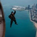 A skydiver in a black jumpsuit and helmet is captured mid-exit from an aircraft, with one arm raised, against a stunning aerial backdrop of Chicago's lakefront. The image shows Lake Michigan, Navy Pier, the Chicago skyline, and the distinctive curved shoreline with sandy beaches. The photo appears to be taken from another aircraft or the same aircraft's exterior, providing a dramatic perspective.