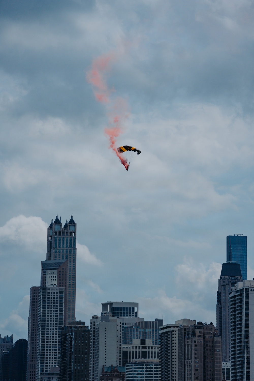 A parachutist with a yellow and black canopy descends over a dense urban skyline, trailing a vivid red/orange smoke plume against a partly cloudy sky. The cityscape in the background features numerous high-rise and skyscraper buildings consistent with a major metropolitan area. This appears to be a demonstration or air show performance over the city.