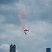 A parachutist with a yellow and black canopy descends over a dense urban skyline, trailing a vivid red/orange smoke plume against a partly cloudy sky. The cityscape in the background features numerous high-rise and skyscraper buildings consistent with a major metropolitan area. This appears to be a demonstration or air show performance over the city.