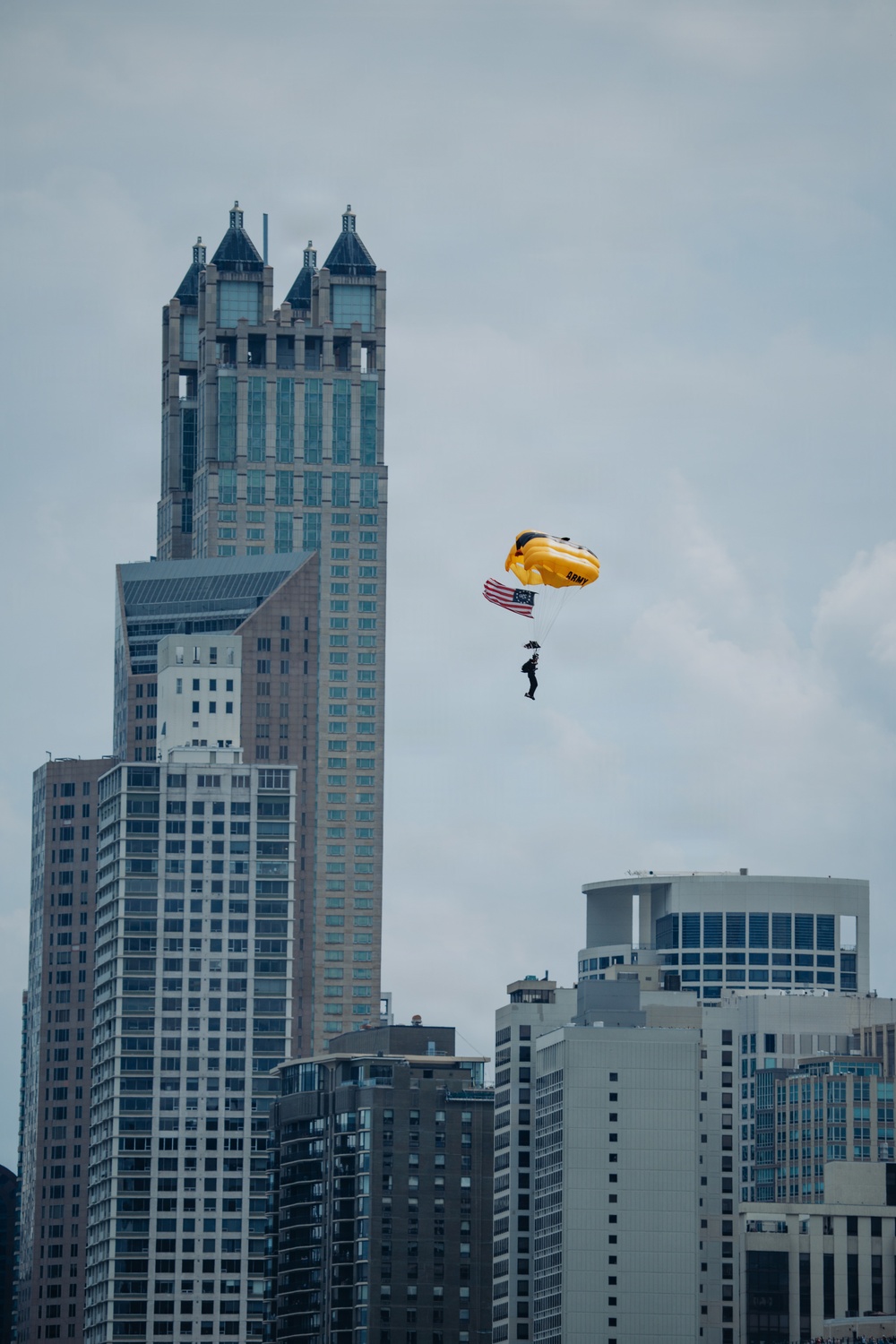 A U.S. Army parachutist descends over a dense urban skyline, carrying an American flag beneath a bright yellow parachute marked 'ARMY'. The backdrop features several prominent skyscrapers, including a distinctive twin-towered building with ornate Gothic-style crown elements. The scene appears to be part of a military demonstration or air show over a major city.