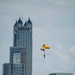 A U.S. Army parachutist descends over a dense urban skyline, carrying an American flag beneath a bright yellow parachute marked 'ARMY'. The backdrop features several prominent skyscrapers, including a distinctive twin-towered building with ornate Gothic-style crown elements. The scene appears to be part of a military demonstration or air show over a major city.