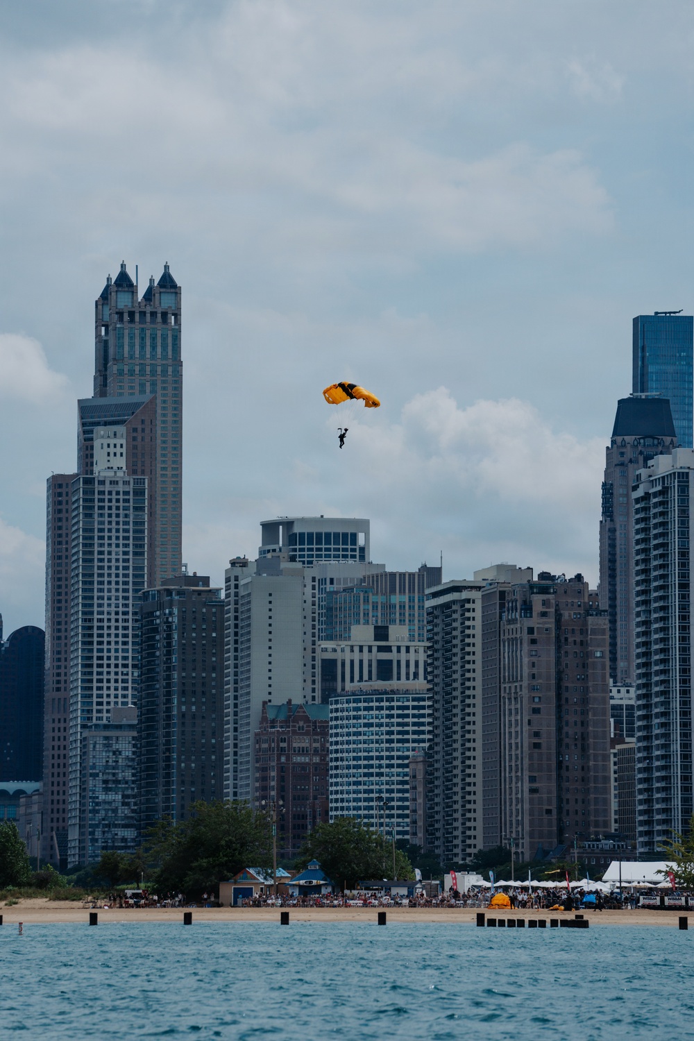 A parachutist with a yellow and black canopy descends over a lakefront beach area with a dense urban skyline in the background. A large crowd is gathered along the sandy shoreline, suggesting a public air show or demonstration event. The scene is photographed from across the water, likely Lake Michigan, with Chicago's distinctive skyline visible.