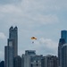 A parachutist with a yellow and black canopy descends over a lakefront beach area with a dense urban skyline in the background. A large crowd is gathered along the sandy shoreline, suggesting a public air show or demonstration event. The scene is photographed from across the water, likely Lake Michigan, with Chicago's distinctive skyline visible.