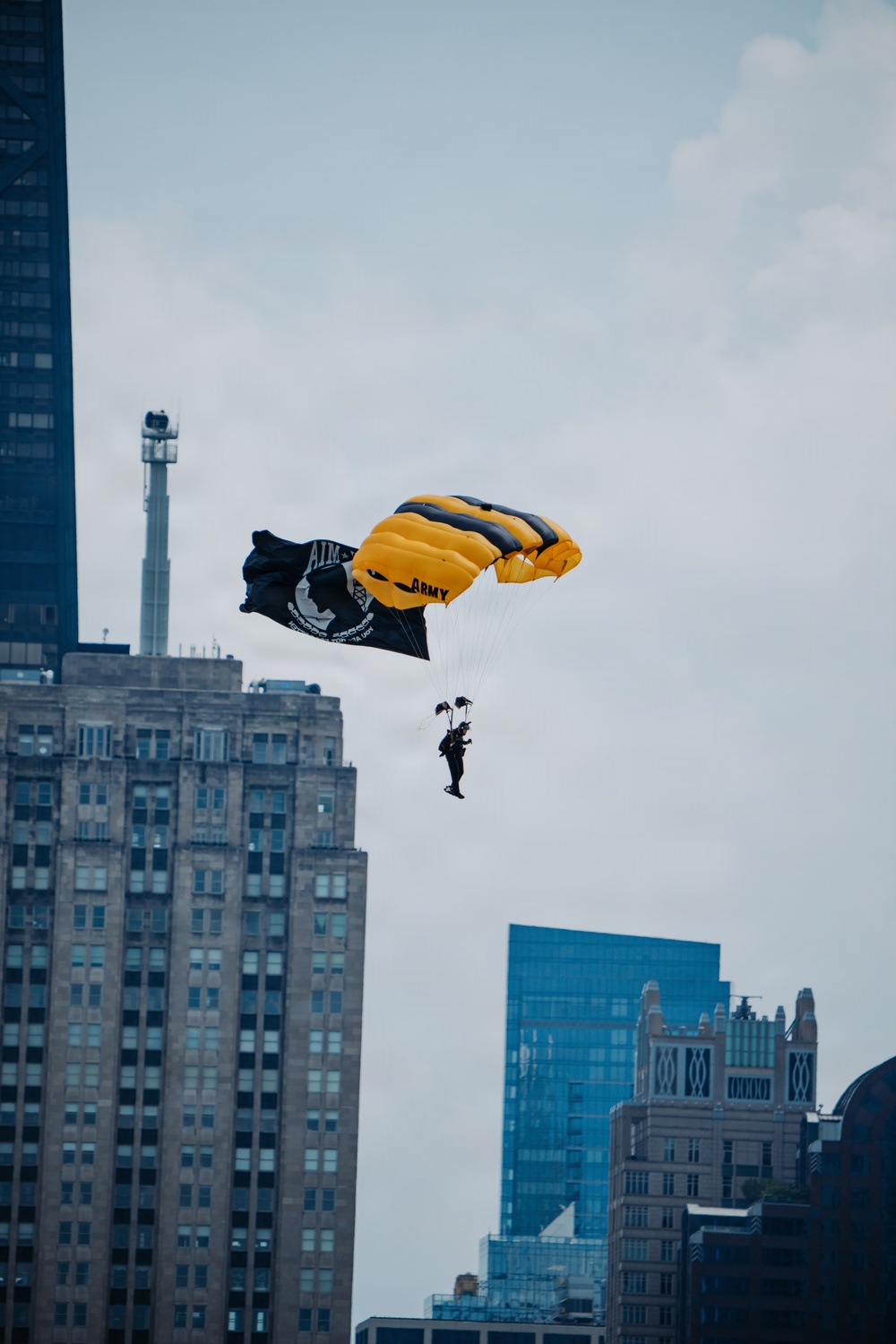 A U.S. Army parachutist descends over an urban skyline with a yellow and black parachute bearing the word 'ARMY'. The jumper is carrying a POW/MIA flag, indicating this is likely a ceremonial or commemorative demonstration jump. The backdrop features recognizable high-rise buildings consistent with a major American city.
