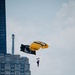A U.S. Army parachutist descends over an urban skyline with a yellow and black parachute bearing the word 'ARMY'. The jumper is carrying a POW/MIA flag, indicating this is likely a ceremonial or commemorative demonstration jump. The backdrop features recognizable high-rise buildings consistent with a major American city.
