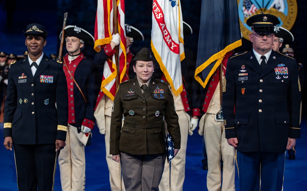 A formal U.S. Army military ceremony featuring three prominent service members standing at attention in the foreground, flanked by a color guard in historical Revolutionary War-era uniforms carrying flags including the U.S. Army flag and American flag. The central figure wears the Army Service Uniform in olive drab/green, while the flanking officers wear the Army Service Uniform in dark blue. The setting appears to be a large indoor venue with ceremonial staging.