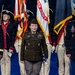A formal U.S. Army military ceremony featuring three prominent service members standing at attention in the foreground, flanked by a color guard in historical Revolutionary War-era uniforms carrying flags including the U.S. Army flag and American flag. The central figure wears the Army Service Uniform in olive drab/green, while the flanking officers wear the Army Service Uniform in dark blue. The setting appears to be a large indoor venue with ceremonial staging.