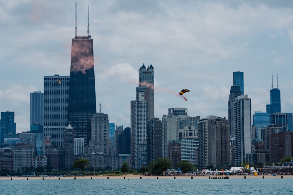 A skydivers or BASE jumper with a yellow and black parachute is visible mid-air against the Chicago skyline, trailing orange/pink smoke. The iconic John Hancock Center (875 North Michigan Avenue) dominates the left side of the frame, with the Willis Tower visible on the far right. The scene appears to be captured from over Lake Michigan during what looks like an air show or demonstration event.