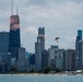 A skydivers or BASE jumper with a yellow and black parachute is visible mid-air against the Chicago skyline, trailing orange/pink smoke. The iconic John Hancock Center (875 North Michigan Avenue) dominates the left side of the frame, with the Willis Tower visible on the far right. The scene appears to be captured from over Lake Michigan during what looks like an air show or demonstration event.