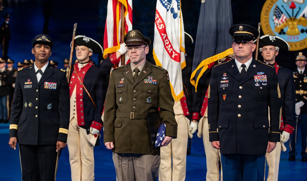 A formal U.S. Army military ceremony featuring three prominent service members standing at attention in the foreground, with a color guard in historical Revolutionary War-era uniforms visible behind them. The central figure holds a folded American flag, suggesting a retirement, award, or memorial ceremony. The backdrop includes U.S. Army flags, the American flag, and what appears to be the U.S. Army seal.