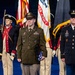 A formal U.S. Army military ceremony featuring three prominent service members standing at attention in the foreground, with a color guard in historical Revolutionary War-era uniforms visible behind them. The central figure holds a folded American flag, suggesting a retirement, award, or memorial ceremony. The backdrop includes U.S. Army flags, the American flag, and what appears to be the U.S. Army seal.