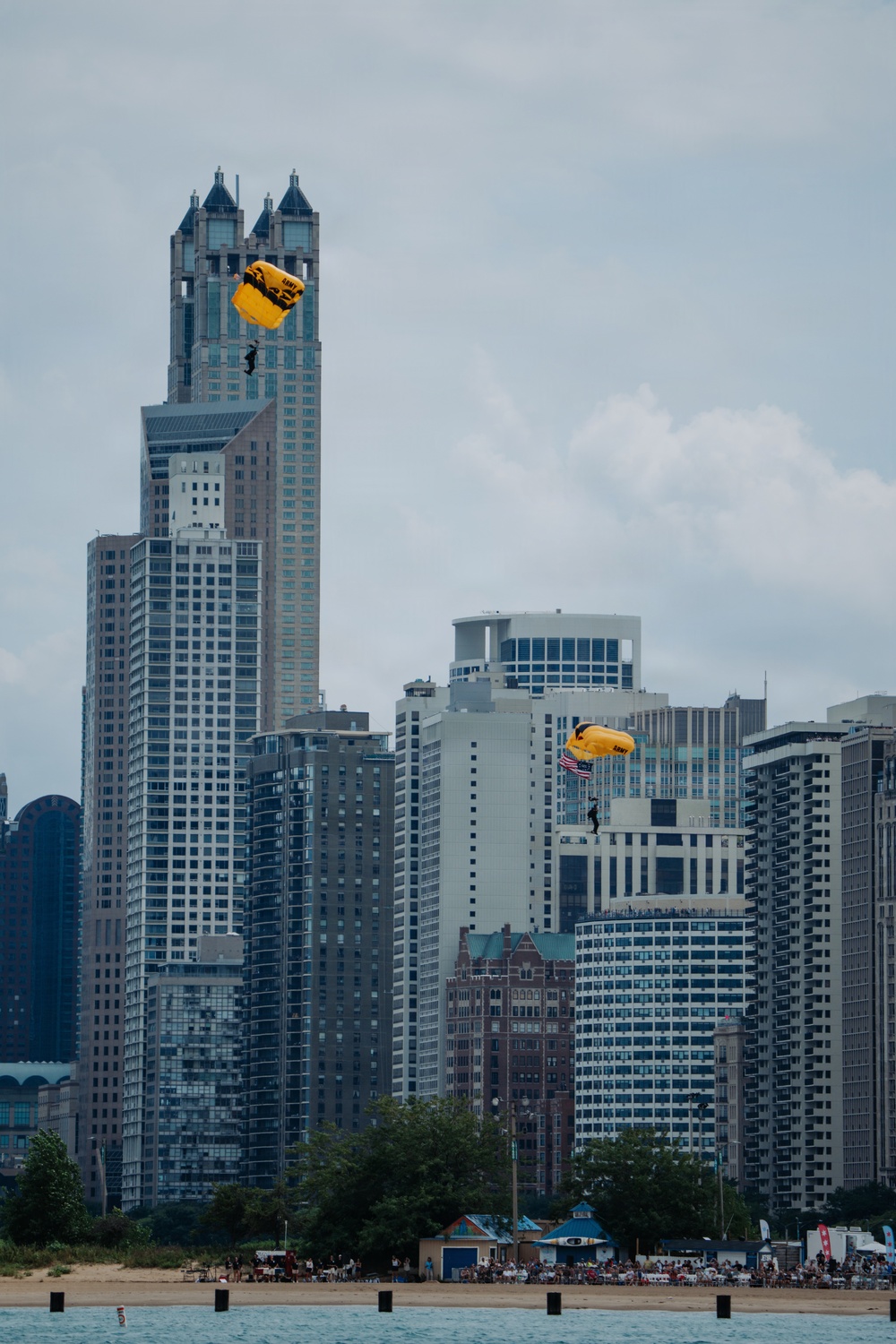 Two U.S. Army parachutists with yellow canopies are descending over a dense urban skyline, identifiable by 'ARMY' text on the parachutes. One parachutist is carrying what appears to be an American flag. A sandy beach with spectators is visible in the foreground, suggesting this is a public airshow or demonstration event.