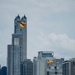 Two U.S. Army parachutists with yellow canopies are descending over a dense urban skyline, identifiable by 'ARMY' text on the parachutes. One parachutist is carrying what appears to be an American flag. A sandy beach with spectators is visible in the foreground, suggesting this is a public airshow or demonstration event.