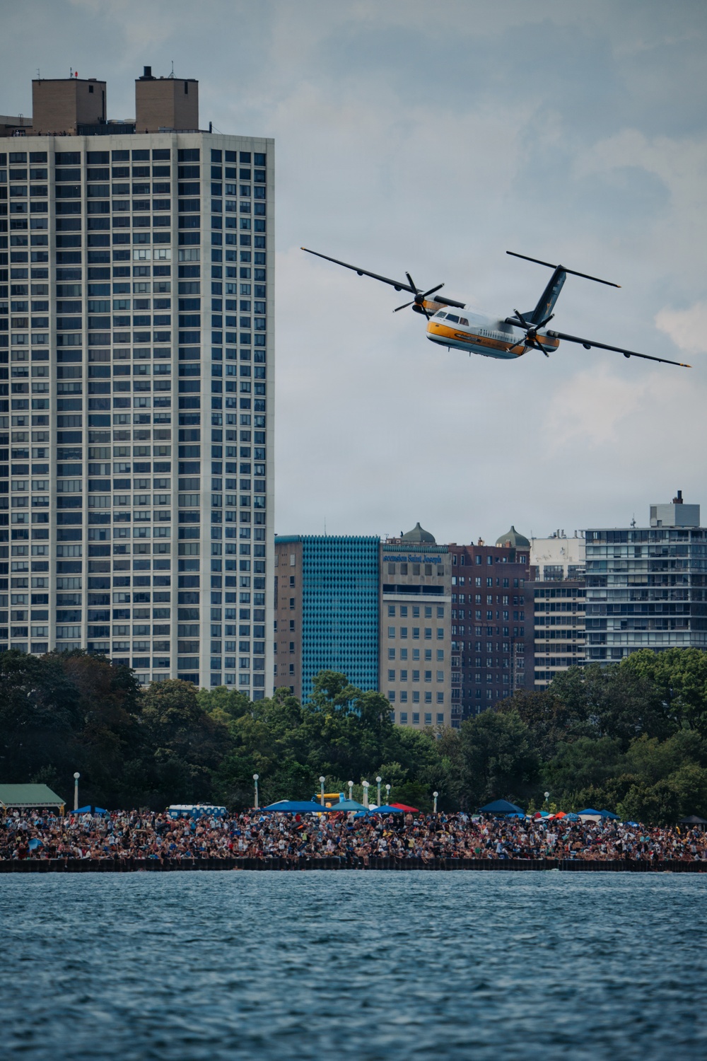 A twin-turboprop aircraft with a white, yellow, and teal color scheme flies low over a waterfront area in front of a large crowd of spectators. The urban skyline features a prominent high-rise residential tower and several mid-rise buildings, one of which bears the text 'Hôpital Saint-Joseph'. The scene appears to be an airshow event viewed from across a body of water.