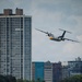 A twin-turboprop aircraft with a white, yellow, and teal color scheme flies low over a waterfront area in front of a large crowd of spectators. The urban skyline features a prominent high-rise residential tower and several mid-rise buildings, one of which bears the text 'Hôpital Saint-Joseph'. The scene appears to be an airshow event viewed from across a body of water.