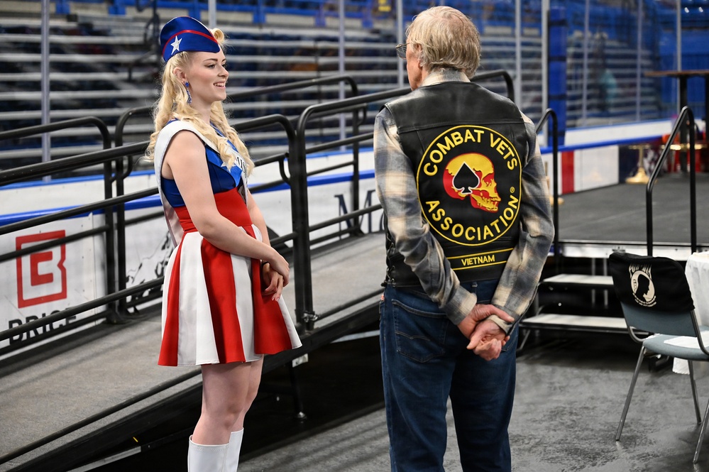 A young woman in a patriotic red, white, and blue majorette or color guard costume with a star-adorned hat is conversing with an older man wearing a leather vest over a plaid shirt. The vest displays a 'Combat Vets Association' patch with a skull and spade symbol, along with a 'Vietnam' rocker patch, identifying him as a Vietnam War veteran. The setting appears to be an indoor ice hockey arena or sports venue.
